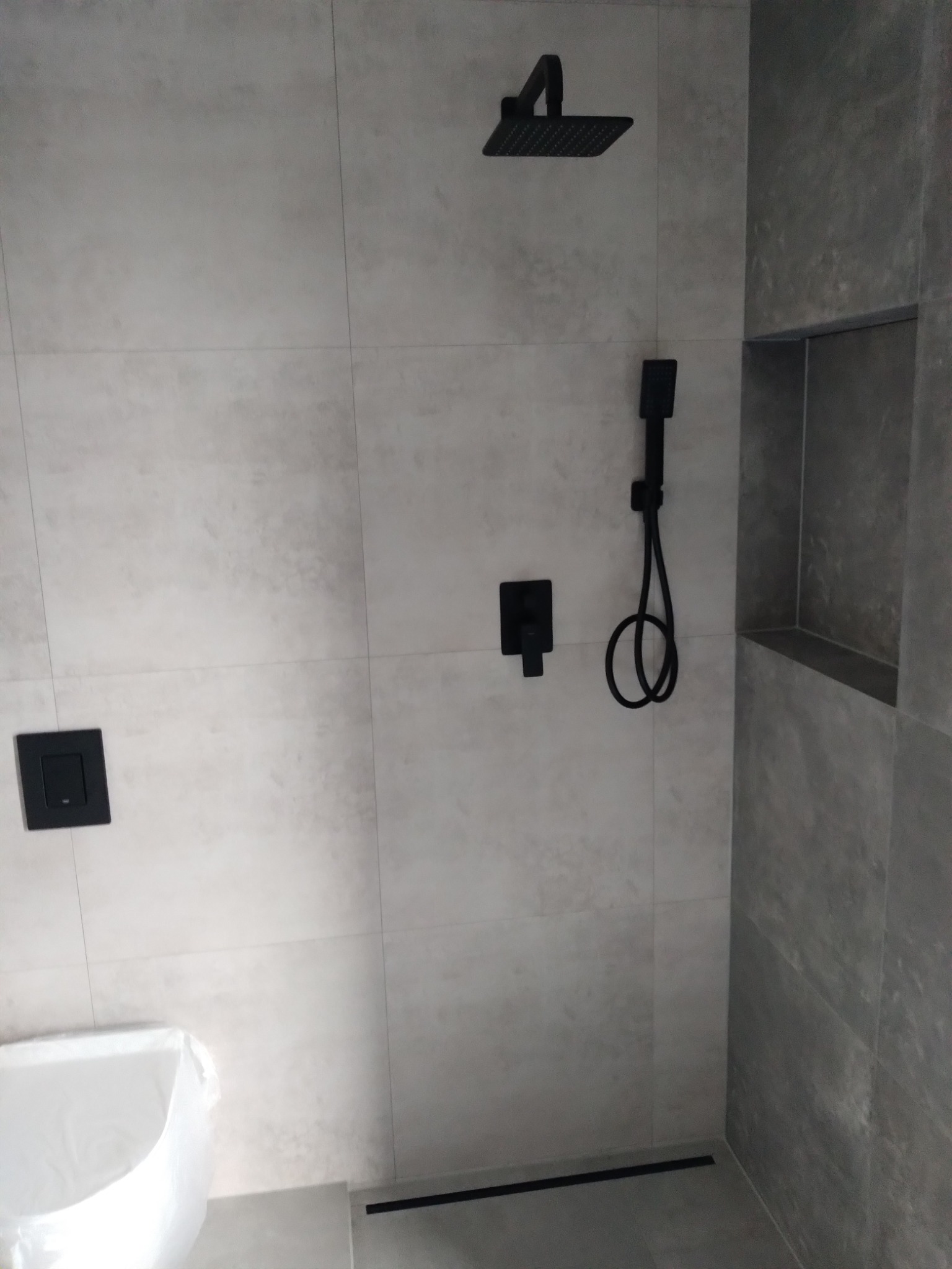 Minimalistyczna kabina prysznicowa z szarymi płytkami i czarną armaturą. Wnęka w ścianie wykończona płytkami. Nowoczesny design i stonowana kolorystyka.