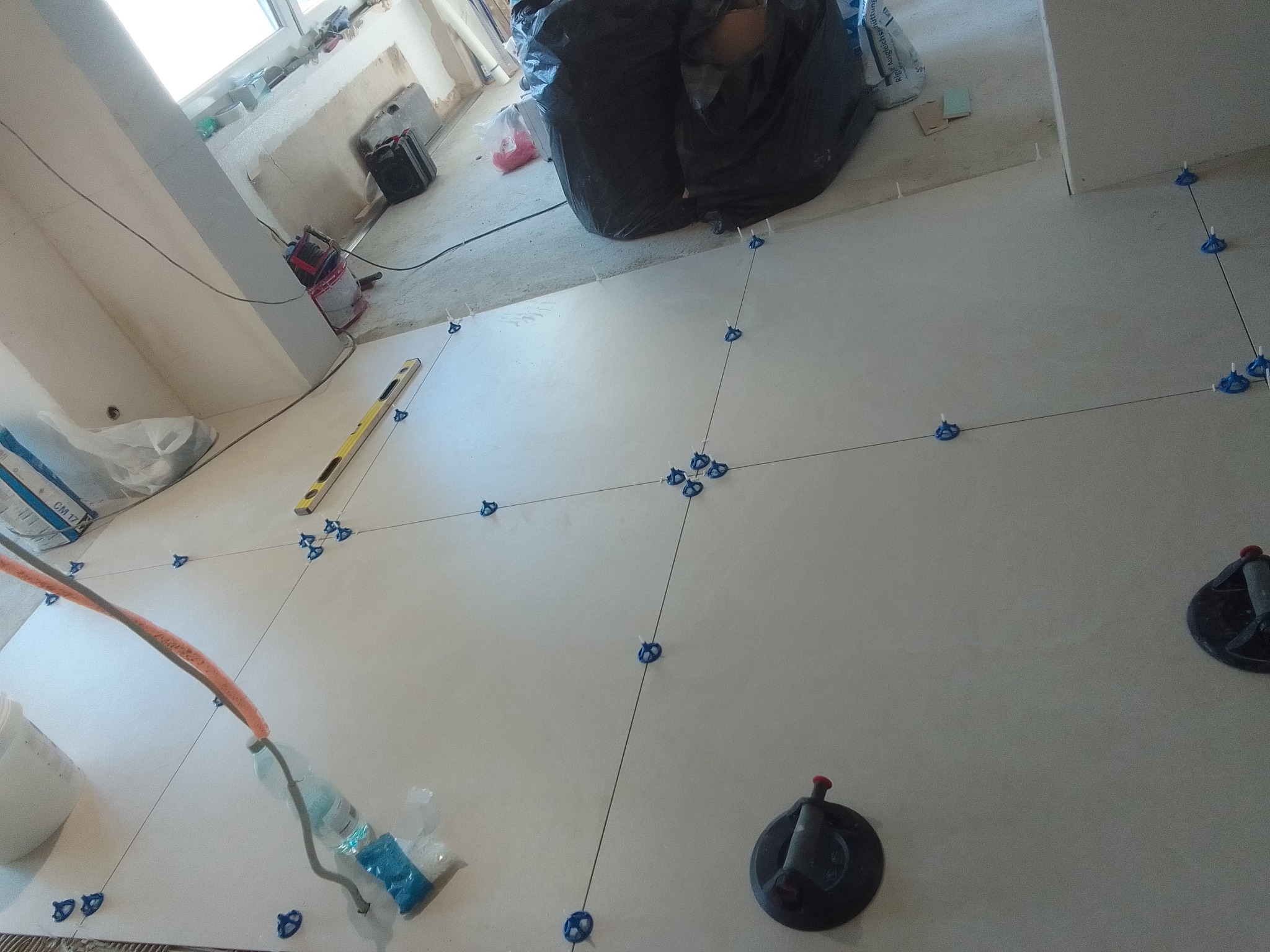 Układanie jasnych płytek podłogowych z użyciem krzyżyków i przyssawek. Widoczny poziomica, worek z klejem i butelka z wodą. Prace glazurnicze w trakcie.