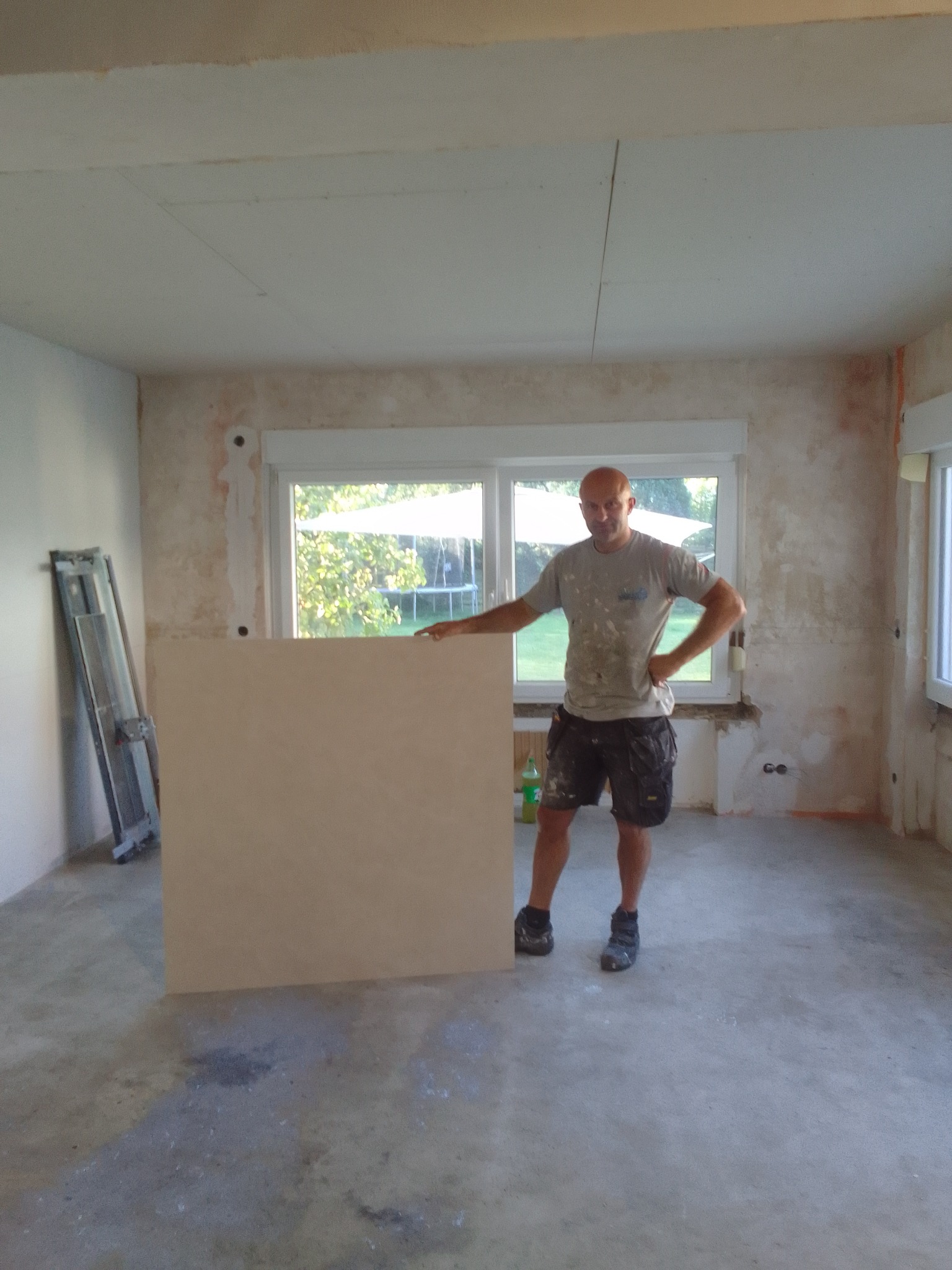 Mężczyzna opiera się o płytę w surowym wnętrzu. Widoczne ślady po remoncie, odsłonięte ściany i sufit z płyt kartonowo-gipsowych. W tle okno z widokiem na zieleń.