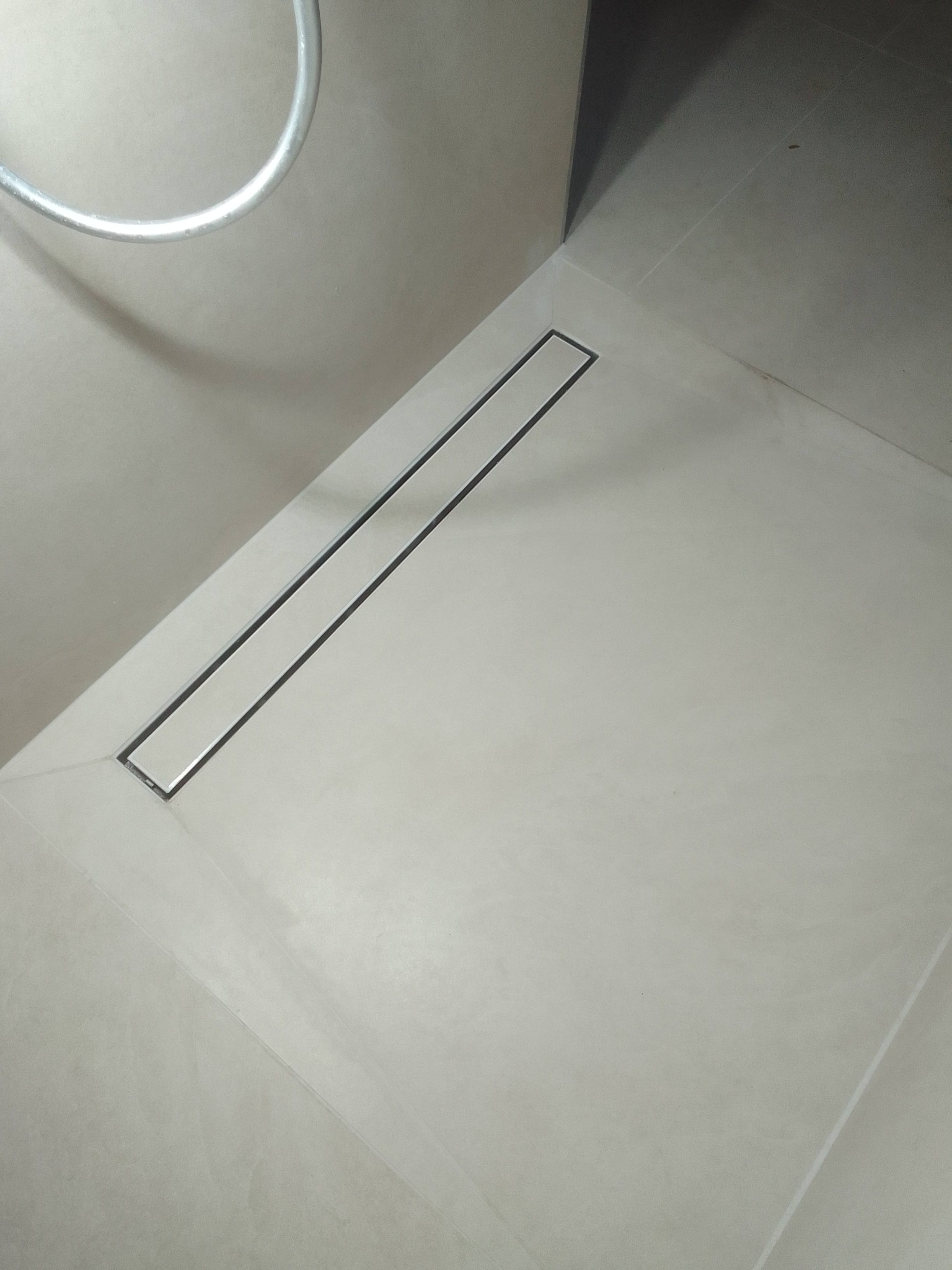 Minimalistyczna łazienka z odpływem liniowym w posadzce, wykończona jasnymi płytkami. Ujęcie z góry, widoczny fragment baterii prysznicowej.