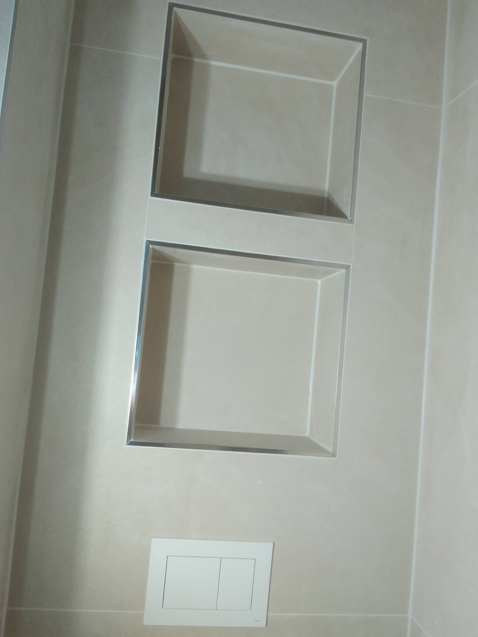 Wykończona łazienka: beżowe płytki z wnękami z chromowanymi ramkami oraz podtynkowy panel WC z dwoma przyciskami. Minimalistyczny design.