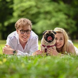 PhotoMagic86 - Uśmiechnięta para z mopsem leży na trawie w parku. Mężczyzna w okularach i zegarku, kobieta z blond włosami. Pies z wywieszonym językiem.