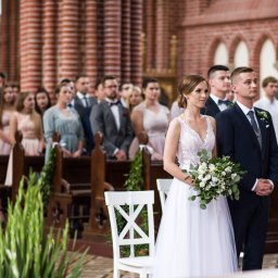 PhotoMagic86 - Para młoda w kościele podczas ceremonii ślubnej, panna młoda trzyma bukiet, goście w tle, ceglane ściany kościoła, oświetlenie naturalne.