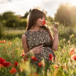 Kobieta w ciąży w kwiecistej sukience, w polu maków, wącha czerwone kwiaty. Delikatne oświetlenie, naturalne tło. Sesja w plenerze, z akcentem na emocje.