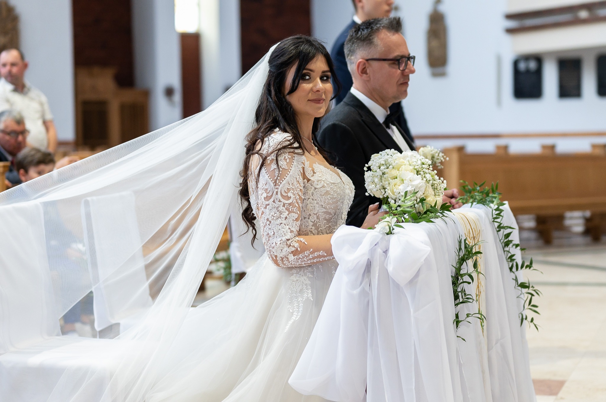 Panna młoda w kościele, w białej sukni z koronkowymi rękawami, spogląda w obiektyw. W tle pan młody i dekoracje kwiatowe na klęczniku.