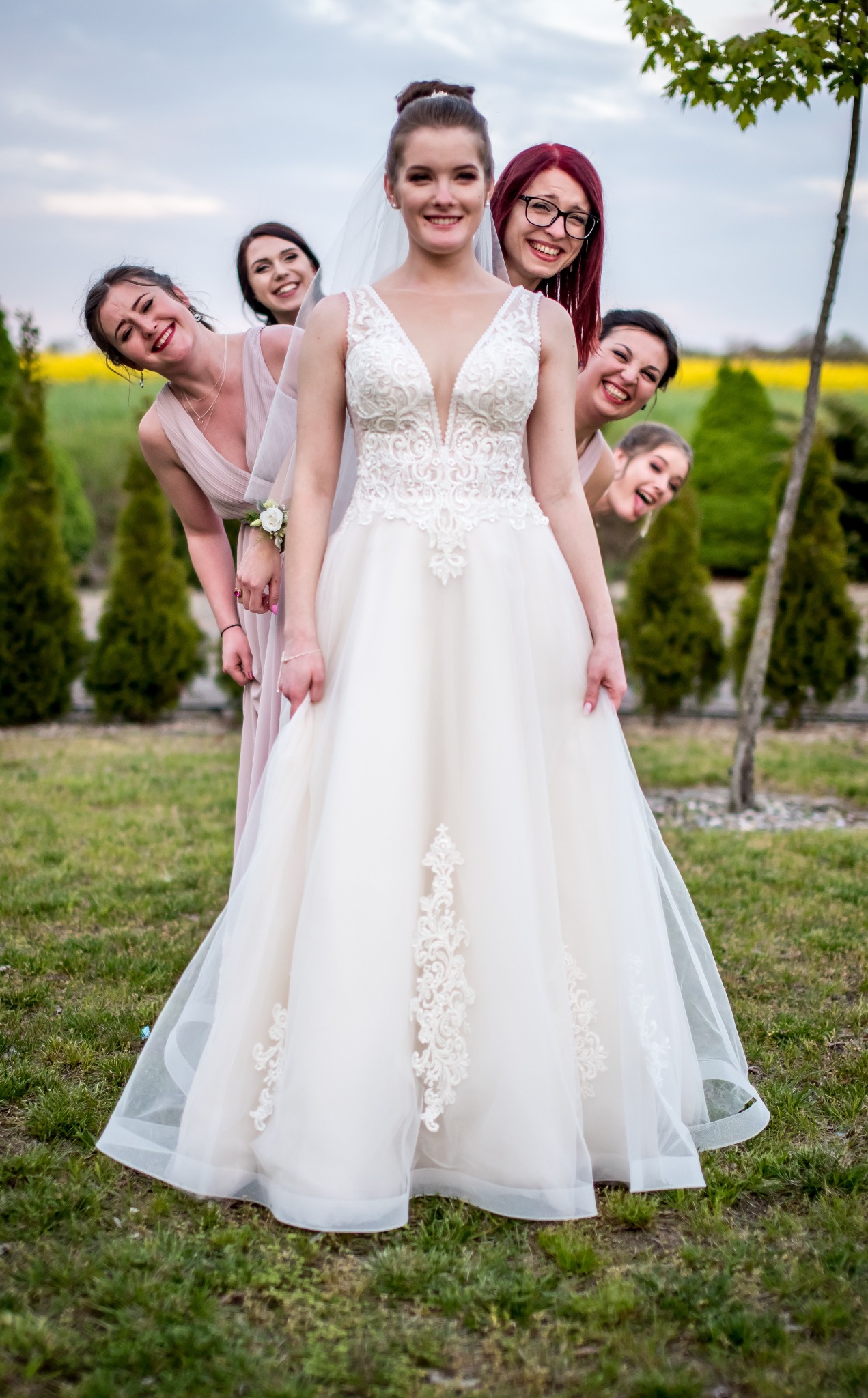 Panna młoda w koronkowej sukni pozuje z uśmiechniętymi druhnami w plenerze we Wrocławiu. W tle drzewa i pole rzepaku, tworzące malowniczy krajobraz.