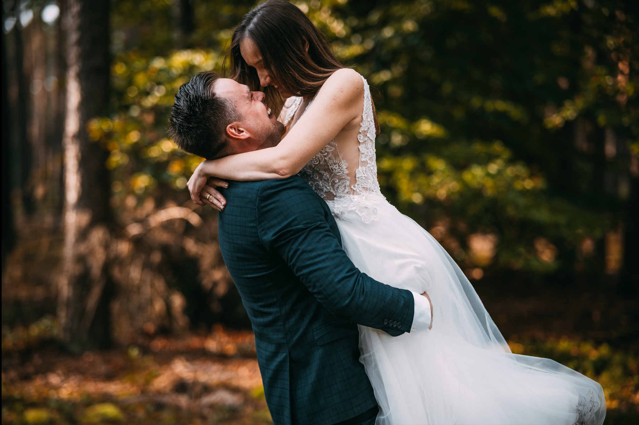 Uśmiechnięty pan młody trzyma pannę młodą w białej sukni na tle drzew w lesie, romantyczna chwila w plenerze, delikatne światło.