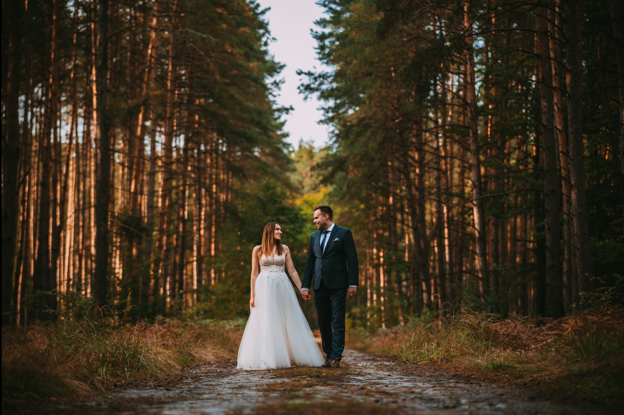 Para młoda, trzymając się za ręce, spaceruje leśną ścieżką. Panna młoda w sukni z koronkową górą, pan młody w ciemnym garniturze. Wysokie drzewa tworzą naturalny tunel.