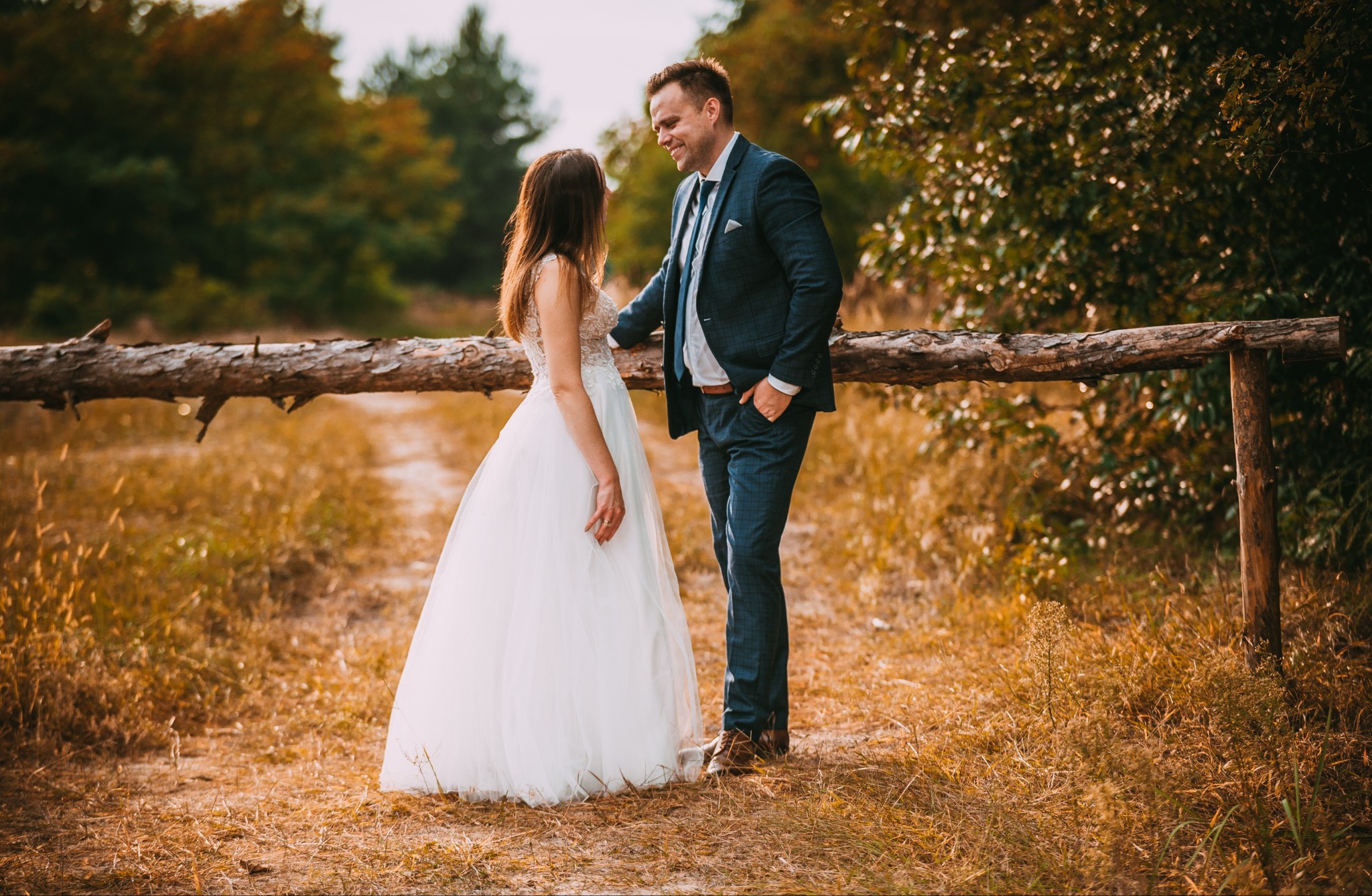 Para młoda w plenerze: panna młoda w białej sukni i pan młody w granatowym garniturze stoją obok drewnianego płotu, patrząc na siebie z uśmiechem, w tle jesienne drzewa.