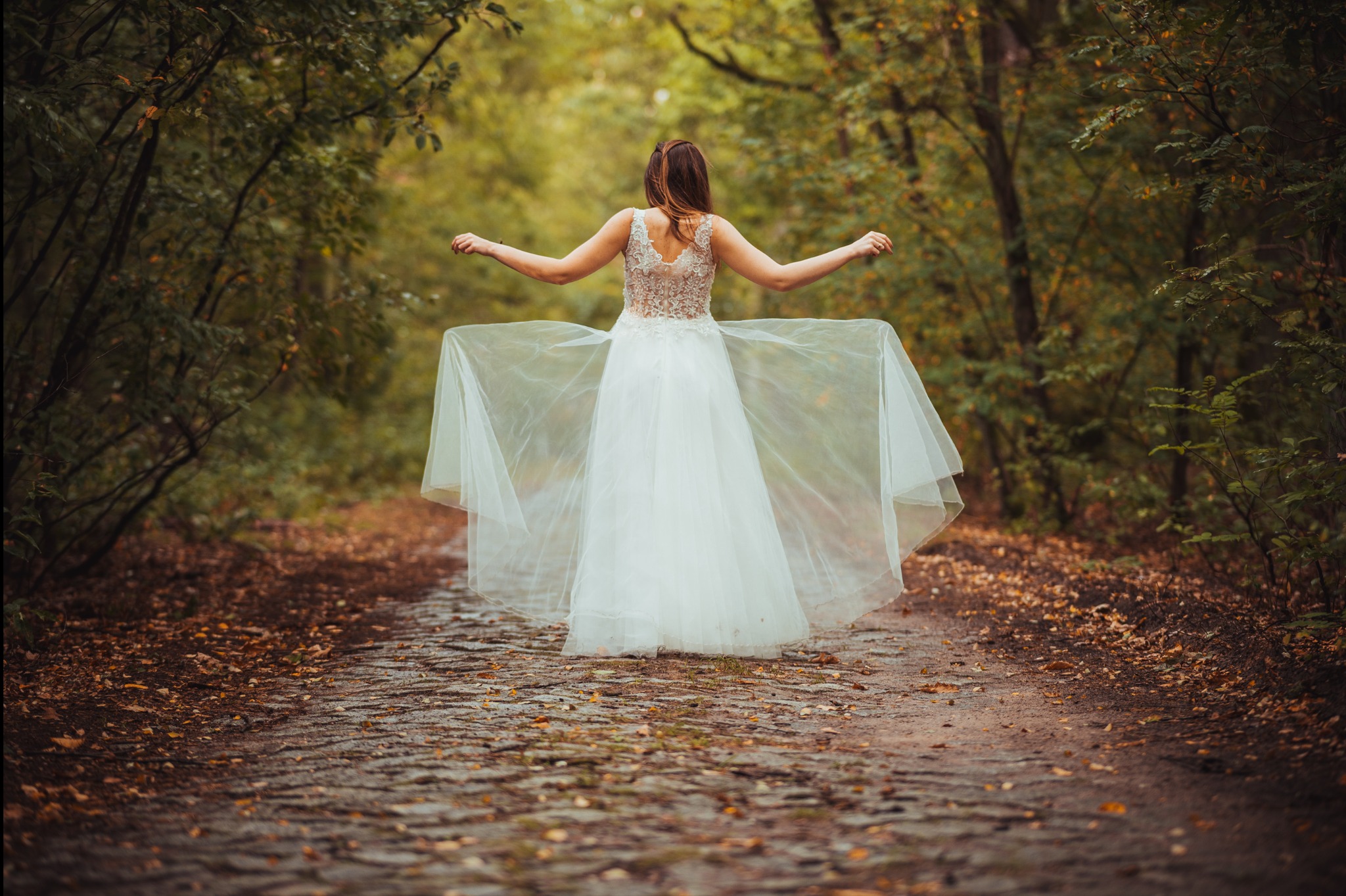 Kobieta w koronkowej sukni ślubnej z rozpostartymi ramionami, idzie leśną ścieżką. Romantyczna sesja w plenerze, widok z tyłu.