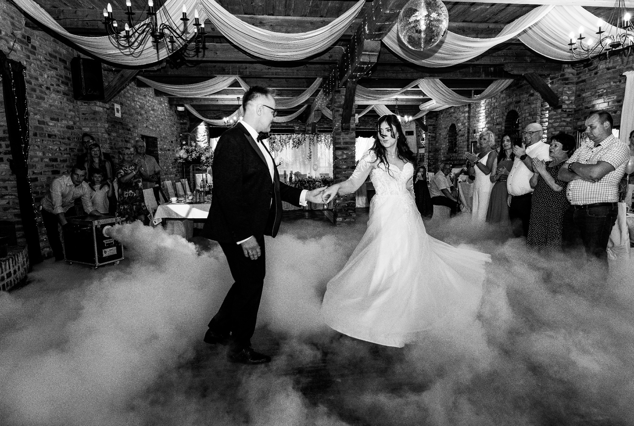 Czarno-białe zdjęcie pary młodej podczas pierwszego tańca w otoczeniu dymu, gości i dekoracji sali weselnej z drewnianymi elementami i żyrandolami.