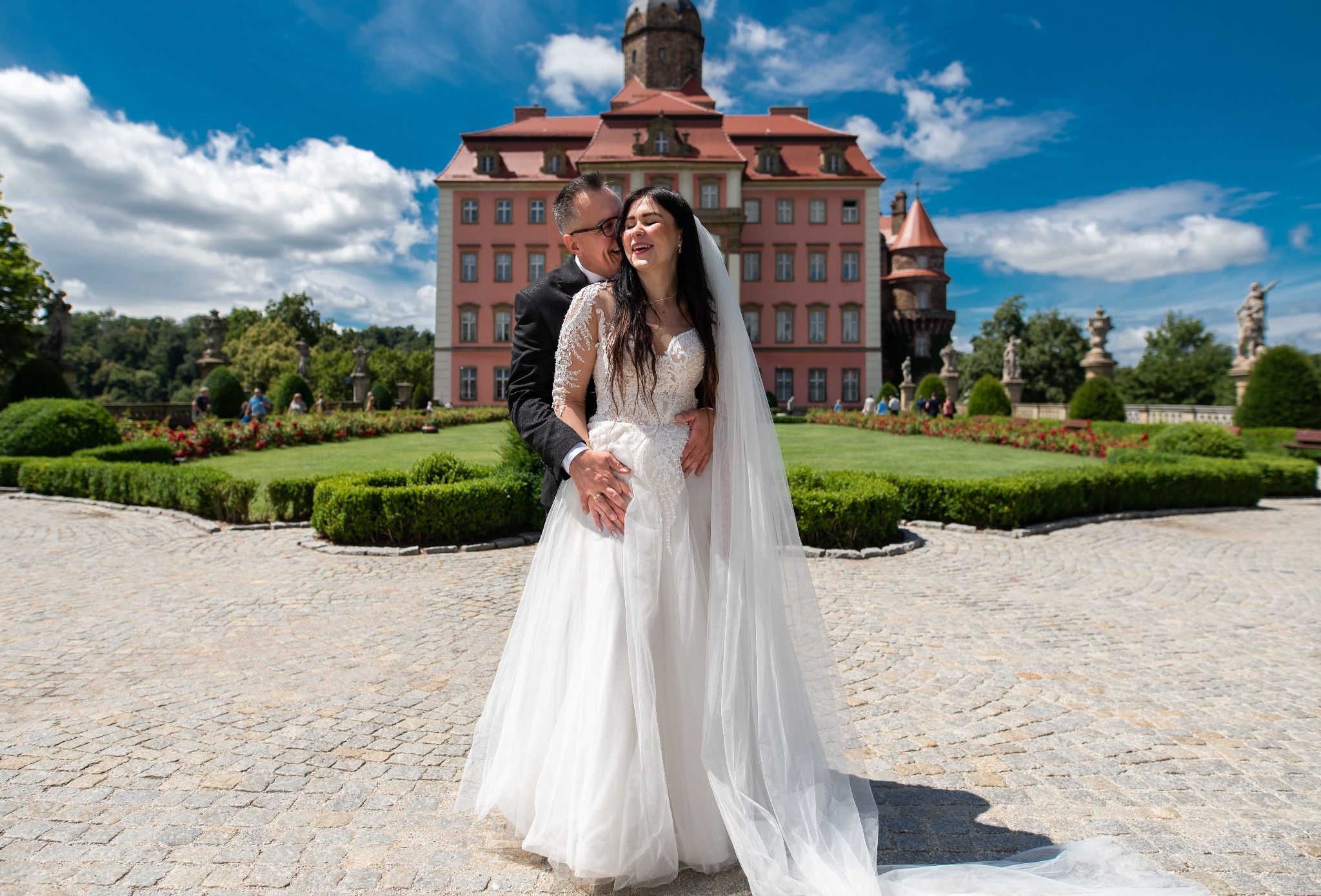 Para młoda w objęciach na tle zamku w Pszczynie. Panna młoda w koronkowej sukni, pan młody w czarnym garniturze. Uśmiechnięci, słoneczny dzień.