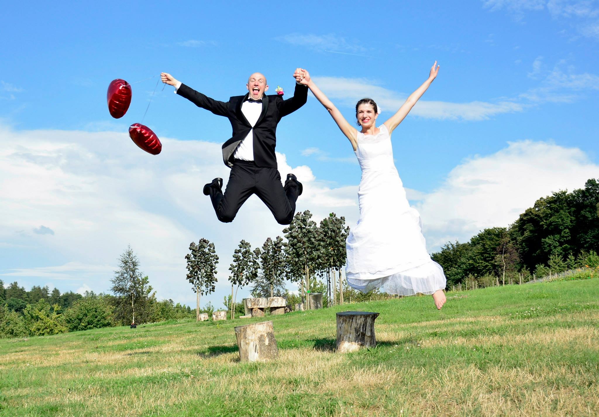 Radosna para młoda w skoku na tle błękitnego nieba i zieleni. Pan młody z balonami, panna młoda w zwiewnej sukni. Nietypowe ujęcie ślubne w plenerze.