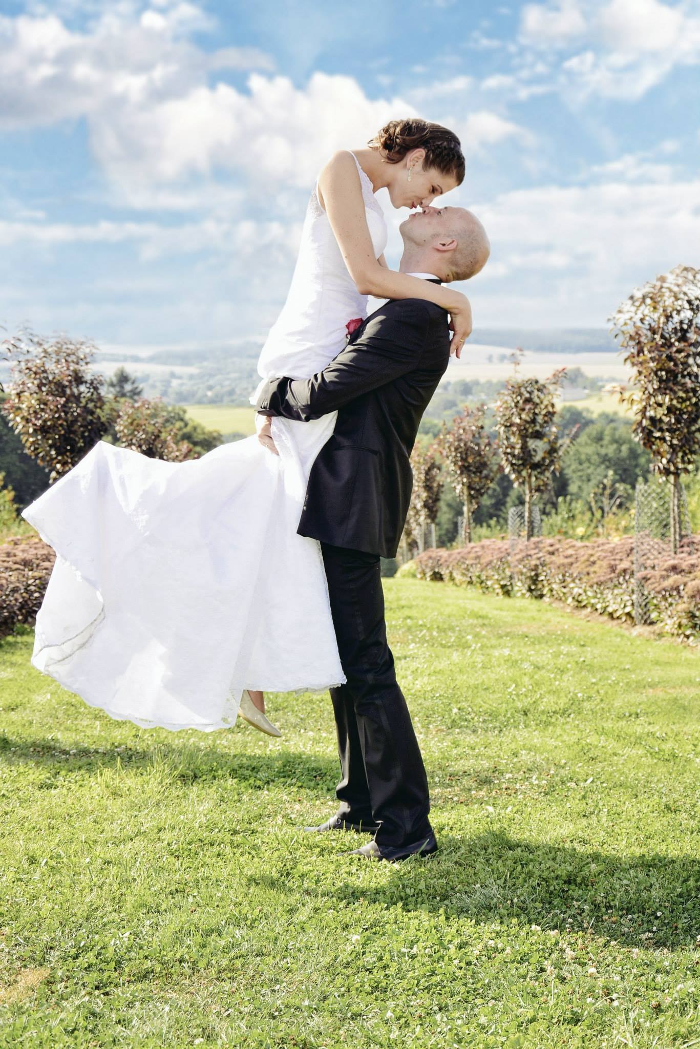 Pan młody unosi pannę młodą w białej sukni, na tle zielonego krajobrazu i błękitnego nieba z chmurami. Romantyczny moment ślubny w plenerze.