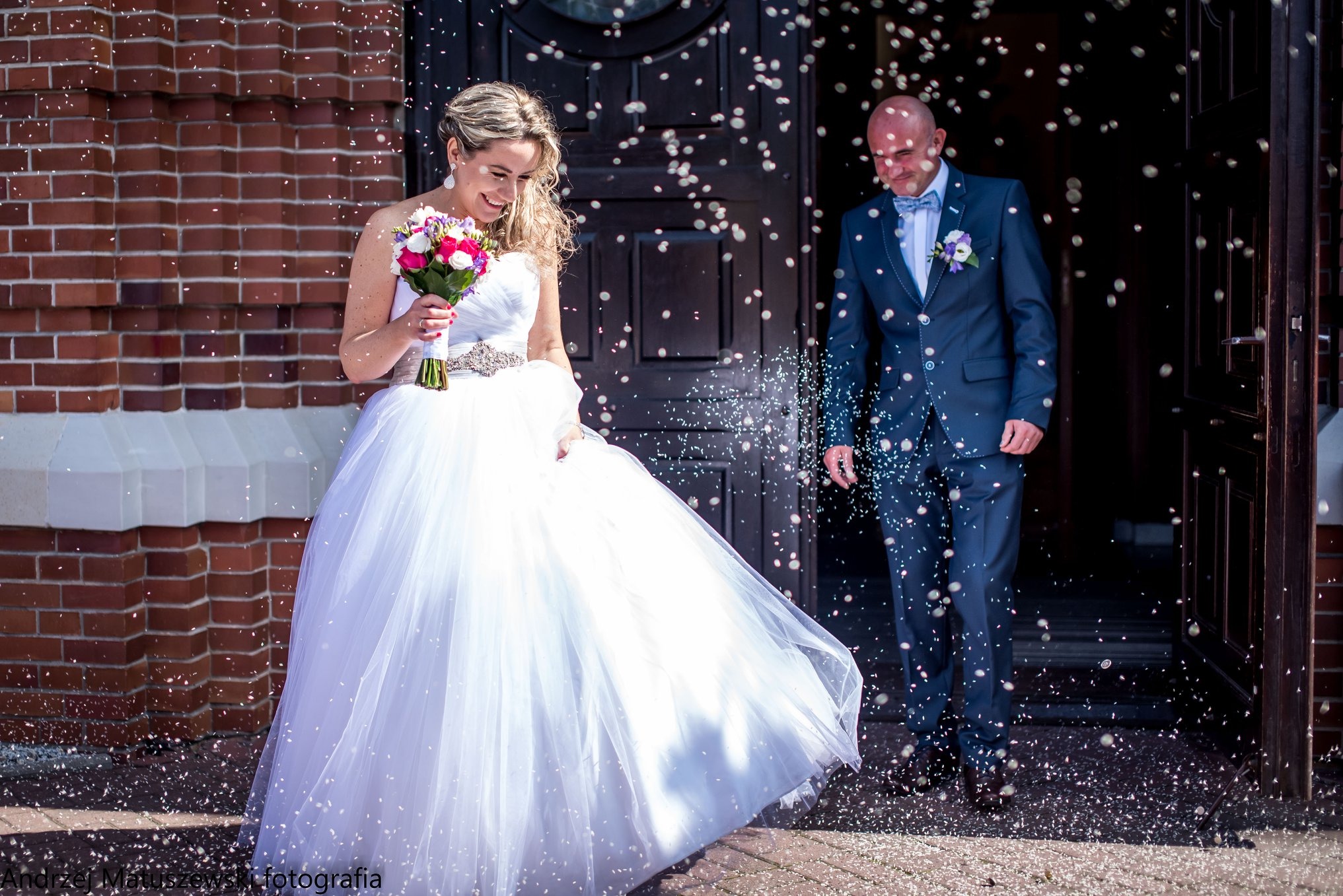 Uśmiechnięta panna młoda z bukietem wychodzi z kościoła, obsypywana konfetti, obok stoi pan młody w granatowym garniturze. Ceglana ściana w tle.