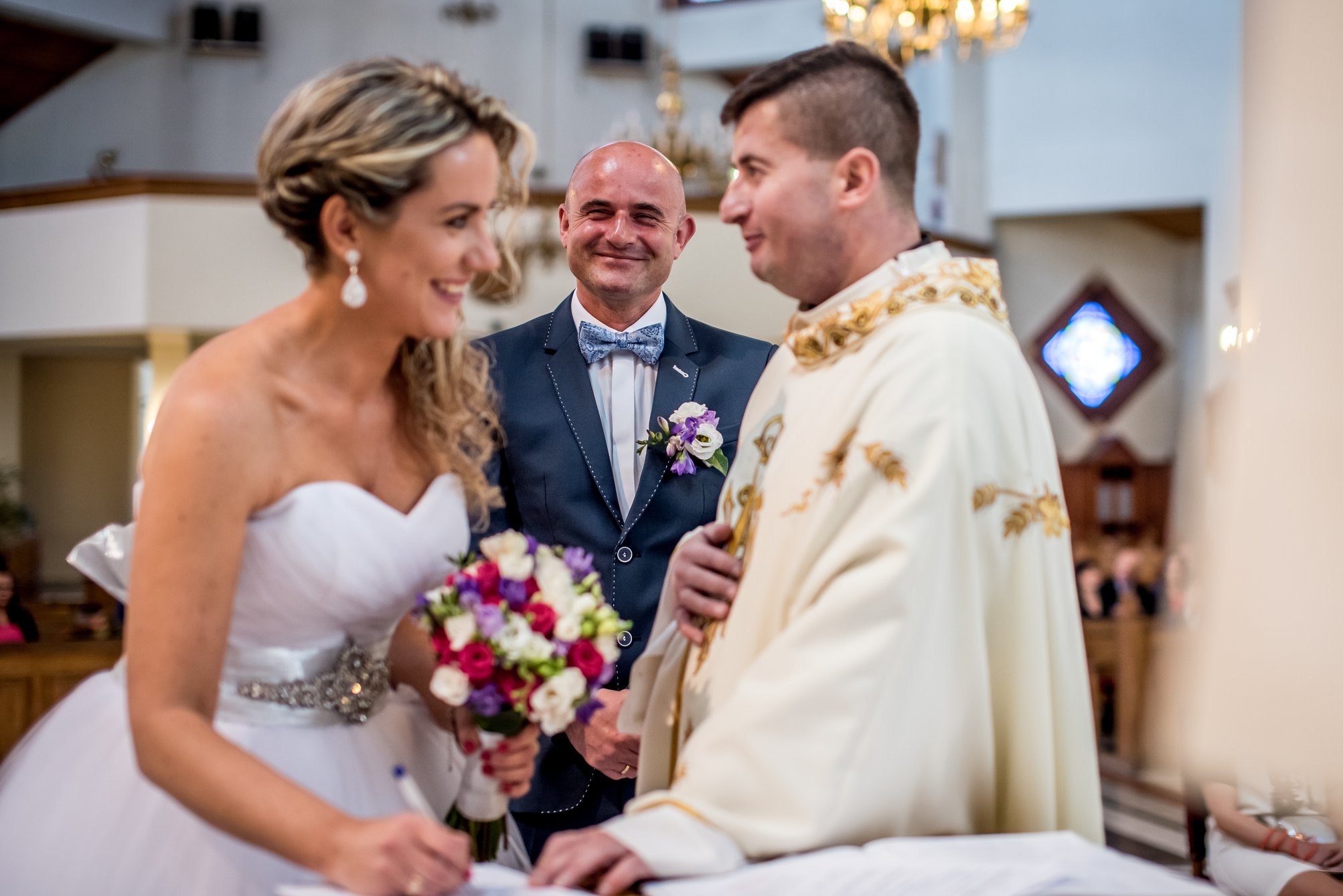 Panna młoda podpisuje dokument podczas ceremonii ślubnej w kościele, z uśmiechniętym mężczyzną w tle i księdzem w bogato zdobionej szacie.