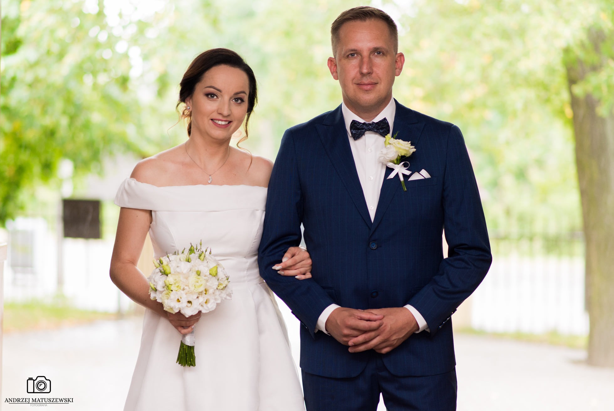 Para młoda: panna młoda w białej sukni z bukietem i pan młody w granatowym garniturze z muszką, pozują na tle zieleni w dniu ślubu.