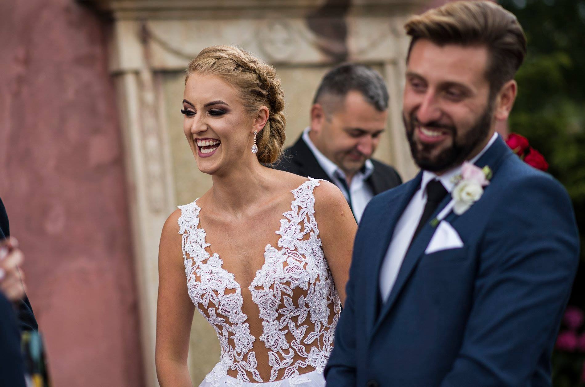 Uśmiechnięta panna młoda w koronkowej sukni i pan młody w granatowym garniturze, w tle goście weselni. Fotografia ślubna z pleneru.