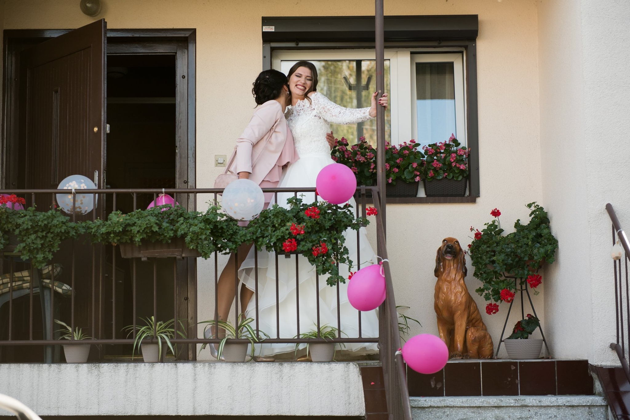 Panna młoda w koronkowej sukni ślubnej, obejmowana przez kobietę w pudrowym różu, na balkonie udekorowanym kwiatami i balonami, z figurą psa. Radosna atmosfera.