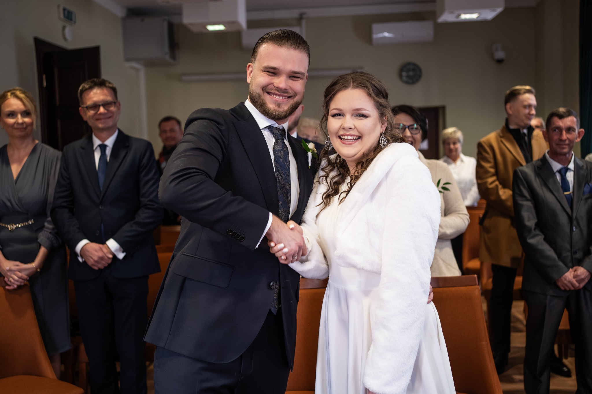Uśmiechnięta para młoda w eleganckich strojach ślubnych ściska dłonie na tle gości podczas ceremonii. Wyraźne emocje i radosna atmosfera.