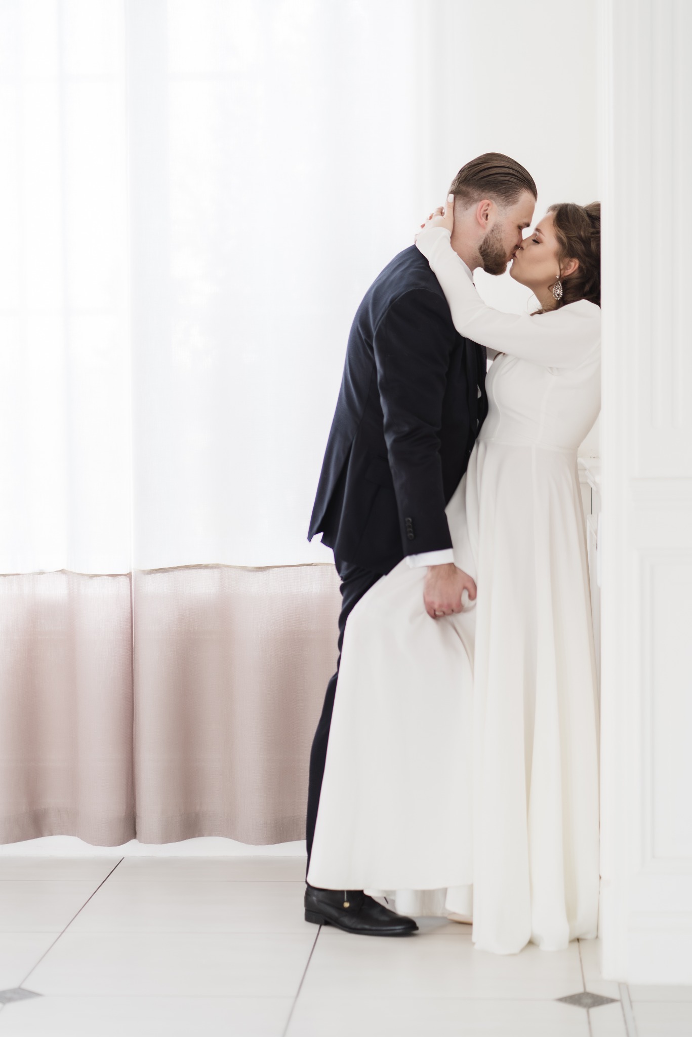 Elegancka para młoda w objęciach, subtelne światło, ona w białej sukni z długim rękawem, on w ciemnym garniturze, delikatny pocałunek w jasnym wnętrzu.