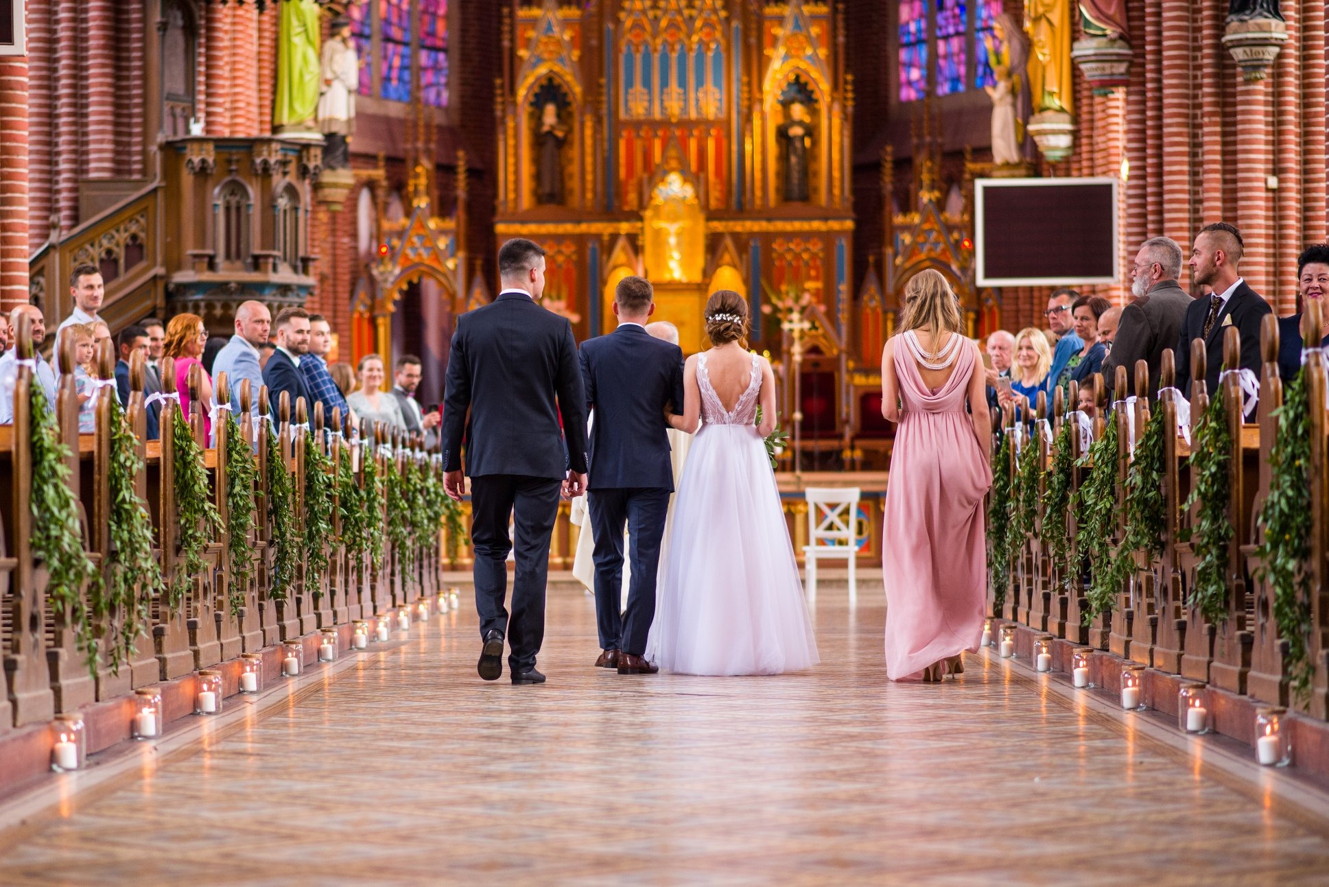 Para młoda prowadzona do ołtarza w kościele. Świece wzdłuż przejścia, goście siedzący w ławkach. Ujęcie z perspektywy od tyłu.