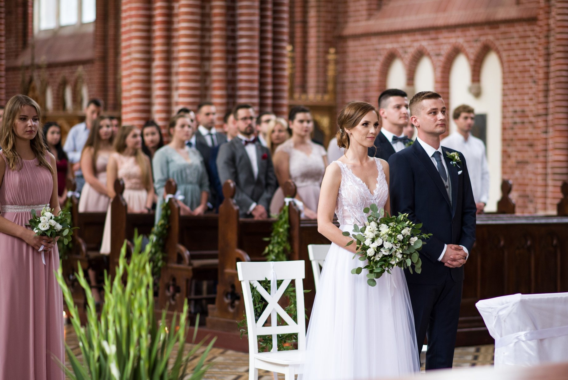 Para młoda w kościele podczas ceremonii ślubnej, panna młoda trzyma bukiet, goście w tle, ceglane ściany kościoła, oświetlenie naturalne.