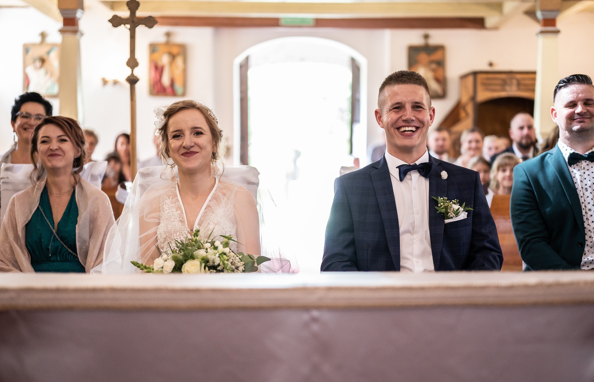Uśmiechnięta para młoda i goście siedzą w kościele podczas ceremonii ślubnej. Pan młody w granatowym garniturze w kratę, panna młoda z bukietem białych róż.