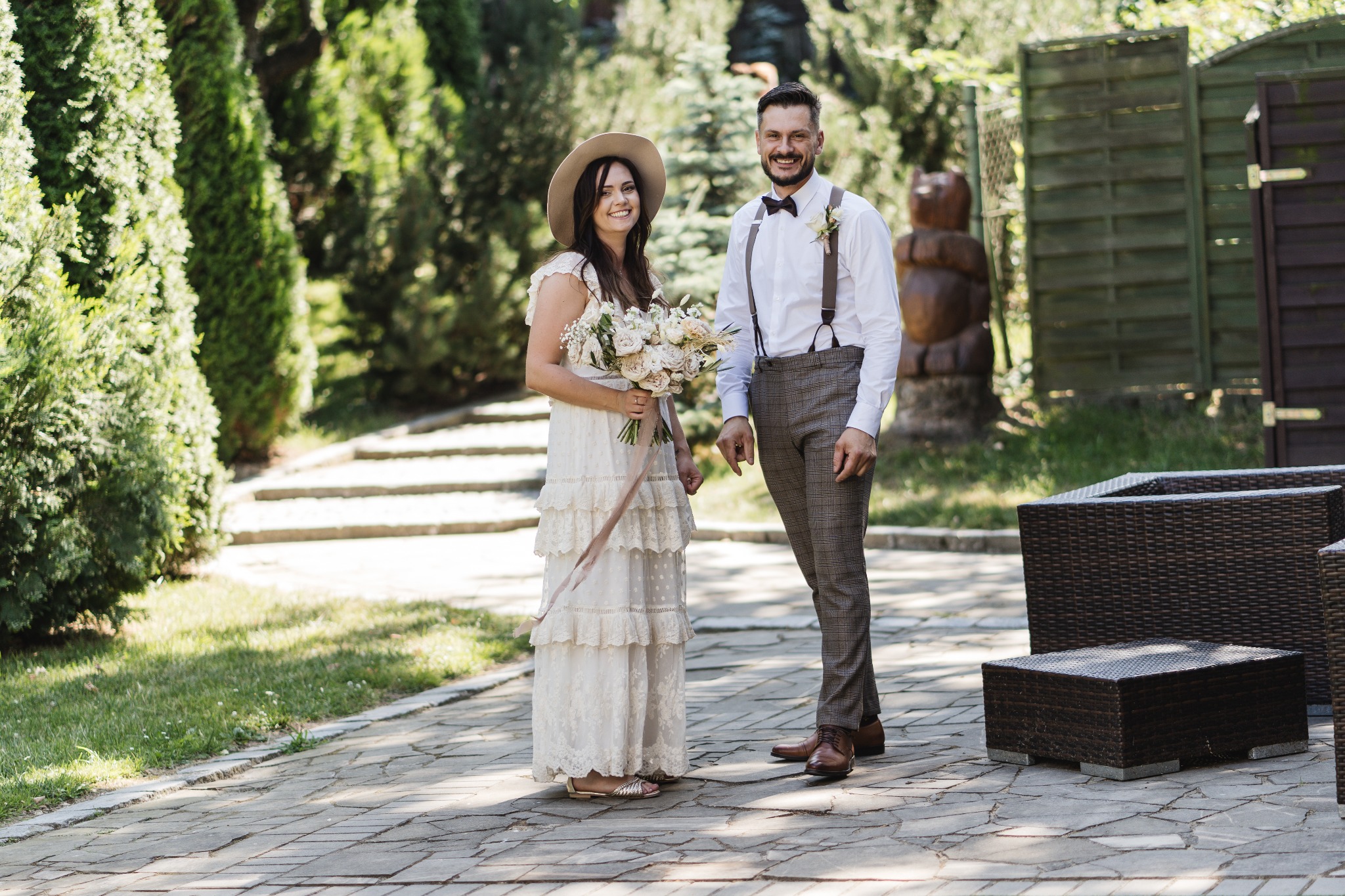 Para młoda w stylu boho na tle zieleni: panna młoda w kapeluszu z bukietem, pan młody w szelkach i muszce. Uśmiechnięci, naturalne światło, plener.