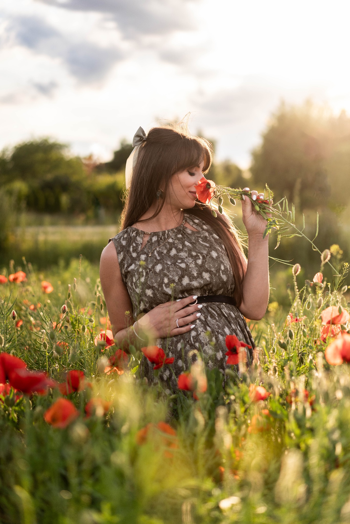 Kobieta w ciąży w kwiecistej sukience, w polu maków, wącha czerwone kwiaty. Delikatne oświetlenie, naturalne tło. Sesja w plenerze, z akcentem na emocje.