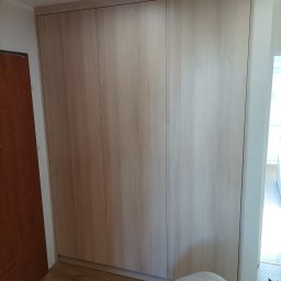 Jolanta Olczyk JR Prestige Meble - Jasna, drewniana szafa wnękowa z trzema drzwiami, zamontowana w pokoju. Na podłodze leżą narzędzia, a osoba w szarej bluzie pracuje nad wykończeniem.