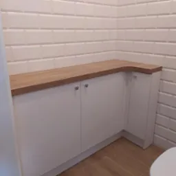 Białe szafki łazienkowe z drewnianym blatem pod umywalką, na tle białych płytek w stylu metro. Chromowane uchwyty, drewniana podłoga.