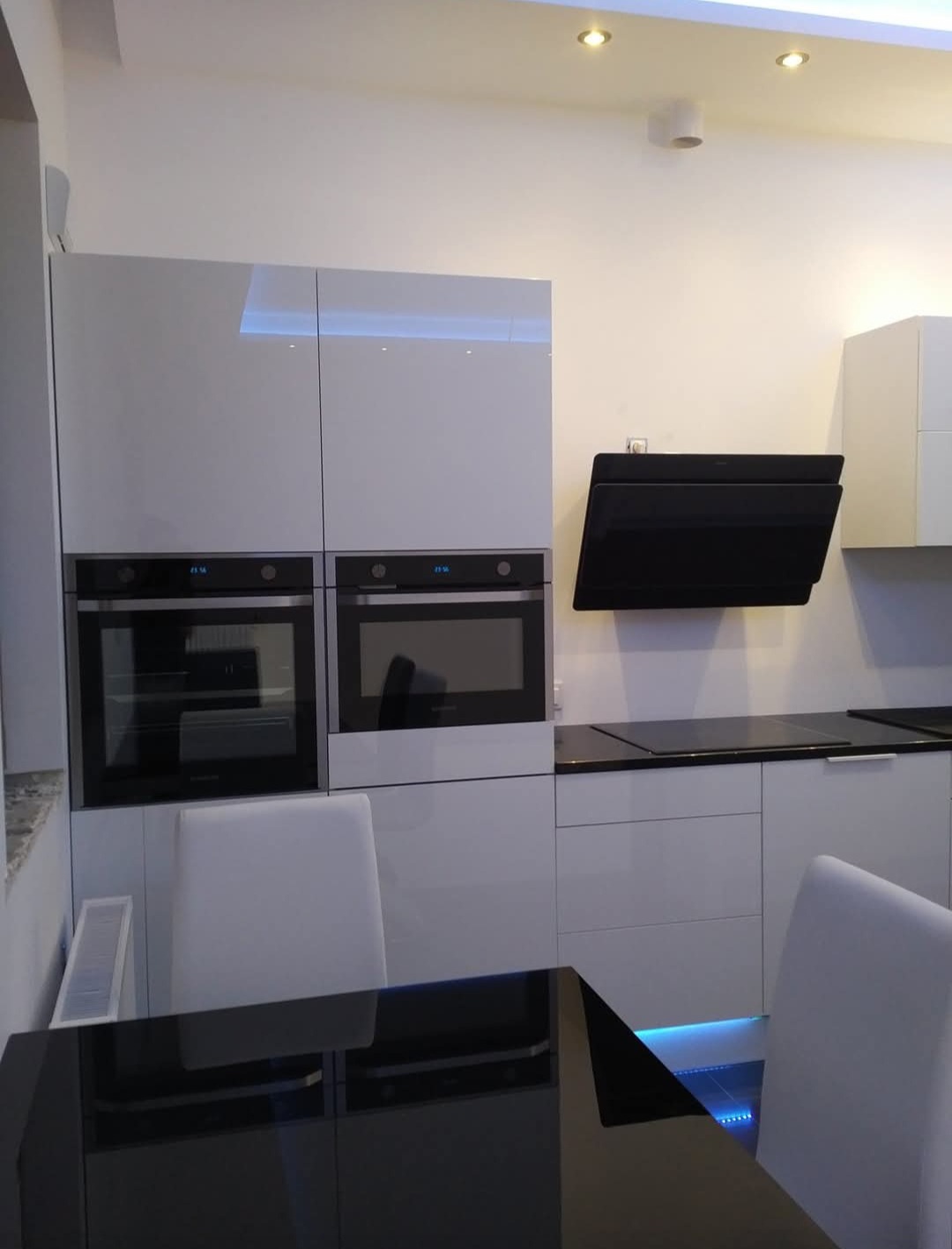 Nowoczesna, biała kuchnia z połyskiem, zabudowana z dwoma piekarnikami i czarnym okapem. Podświetlenie LED pod szafkami dodaje elegancji.