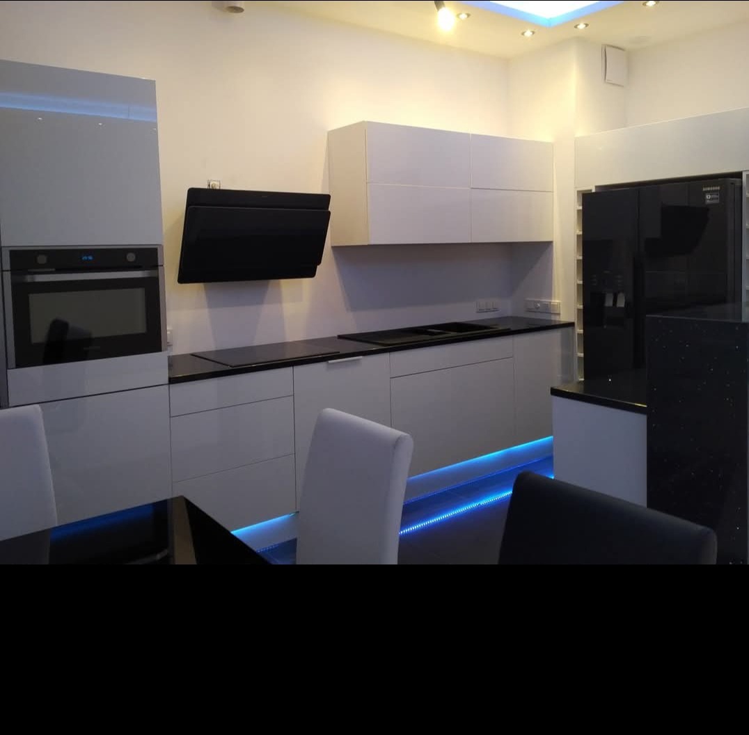 Nowoczesna, biała kuchnia z czarnymi blatami i podświetleniem LED. Na ścianie wisi czarny okap. W tle widoczna czarna lodówka.