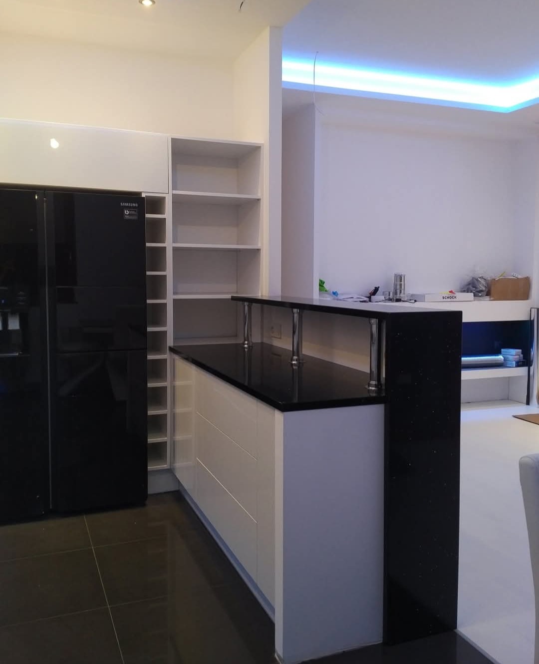 Biała kuchnia z czarnym blatem i lodówką Samsung, minimalistyczny design, podświetlenie LED sufitu, ciemna podłoga, nowoczesne wykończenie, otwarte półki, wysoki połysk.