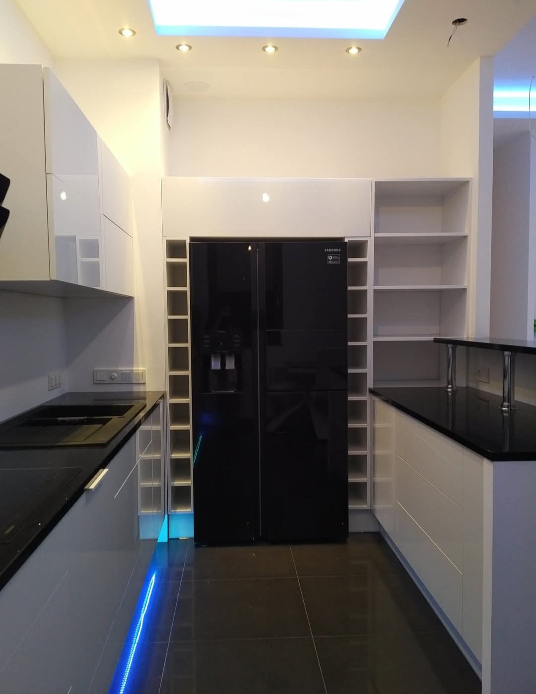Nowoczesna kuchnia z zabudową na wymiar: białe szafki z czarnym blatem, lodówka side-by-side w obudowie z półkami, podświetlenie LED pod szafkami.