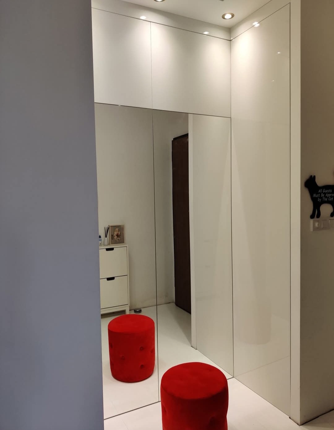 Biała szafa wnękowa z lustrzanymi drzwiami, widoczne dwa czerwone pufy i dekoracja ścienna z motywem kota. Oświetlenie sufitowe podkreśla nowoczesny design.