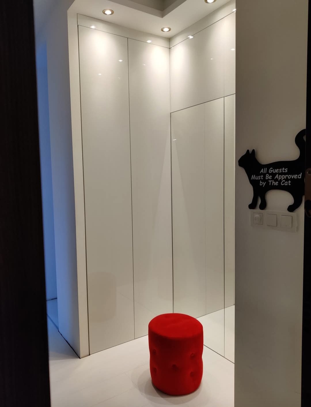 Biała szafa wnękowa z lustrzanymi drzwiami, oświetleniem punktowym i czerwonym pufem. Na ścianie dekoracja z kotem i napisem.