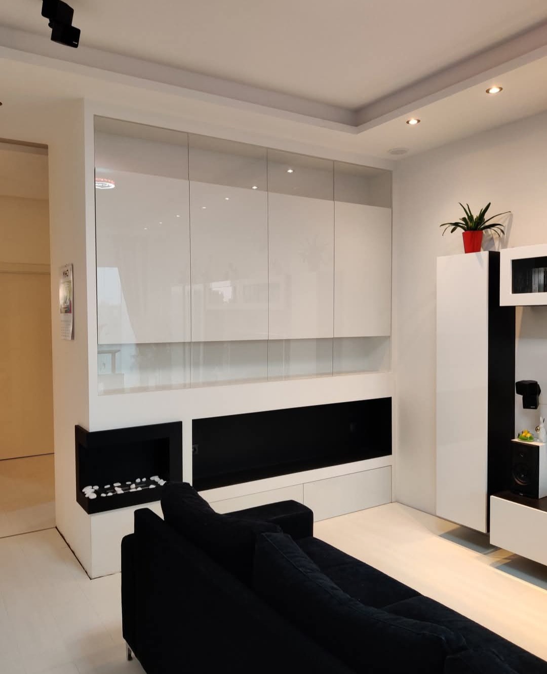 Biała szafa wnękowa z połyskiem, wkomponowana w ścianę salonu z czarnym kominkiem i elementami dekoracyjnymi. Minimalistyczny design i nowoczesne oświetlenie.