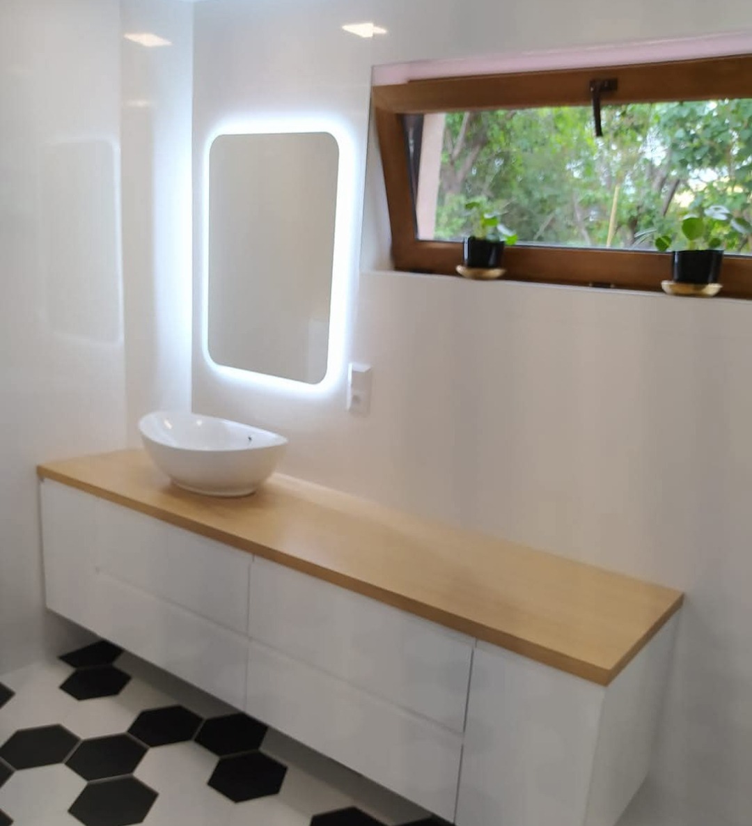 Nowoczesna łazienka z szafką pod umywalkę na wymiar, białe fronty, drewniany blat, podświetlane lustro i heksagonalne płytki podłogowe. Widok na zieleń za oknem.