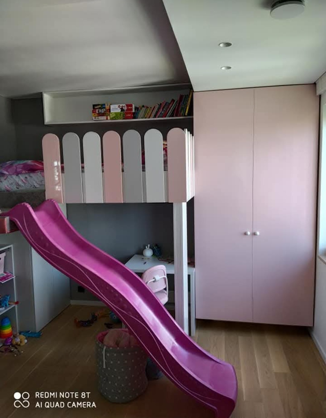 Różowy pokój dziecięcy z łóżkiem na antresoli, zjeżdżalnią i dopasowaną szafą wnękową. Nad łóżkiem półka z książkami, pod spodem biurko.