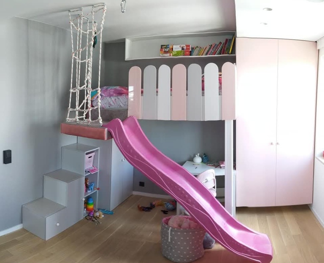 Pokój dziecięcy z łóżkiem na antresoli, różową zjeżdżalnią, siatką do wspinaczki i schodami z szufladami. Zabudowa meblowa z szafą i półkami.