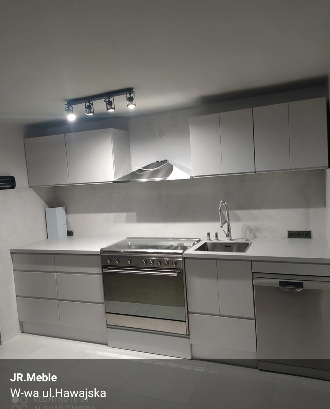 Minimalistyczna, jasnoszara kuchnia z zabudową na wymiar. Widoczne szafki górne i dolne, płyta grzewcza, zlew i oświetlenie punktowe. Nowoczesny design.