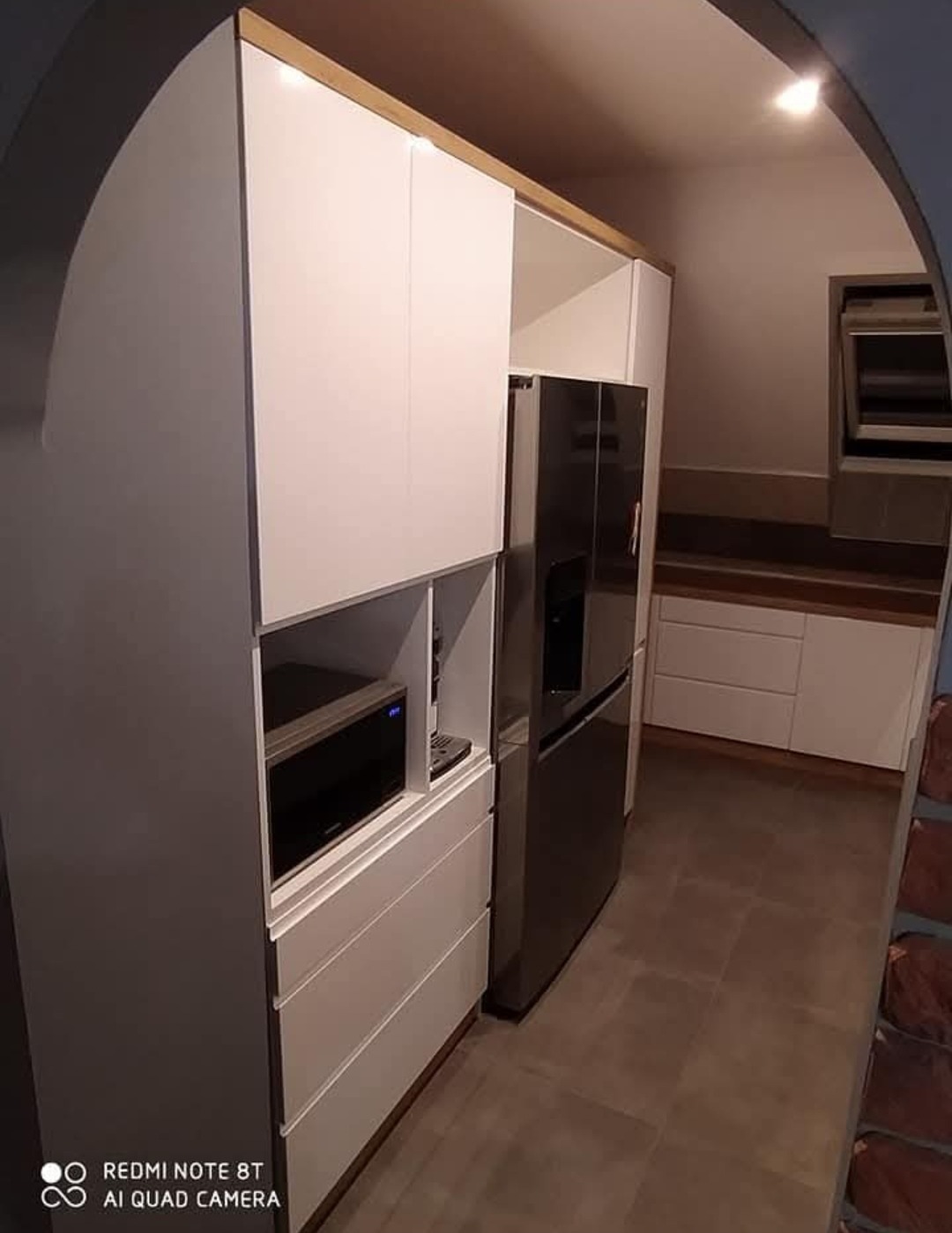 Biała zabudowa kuchenna z drewnianymi akcentami, z lodówką i mikrofalówką, widoczna z perspektywy łuku. Nowoczesny design, minimalistyczny styl.