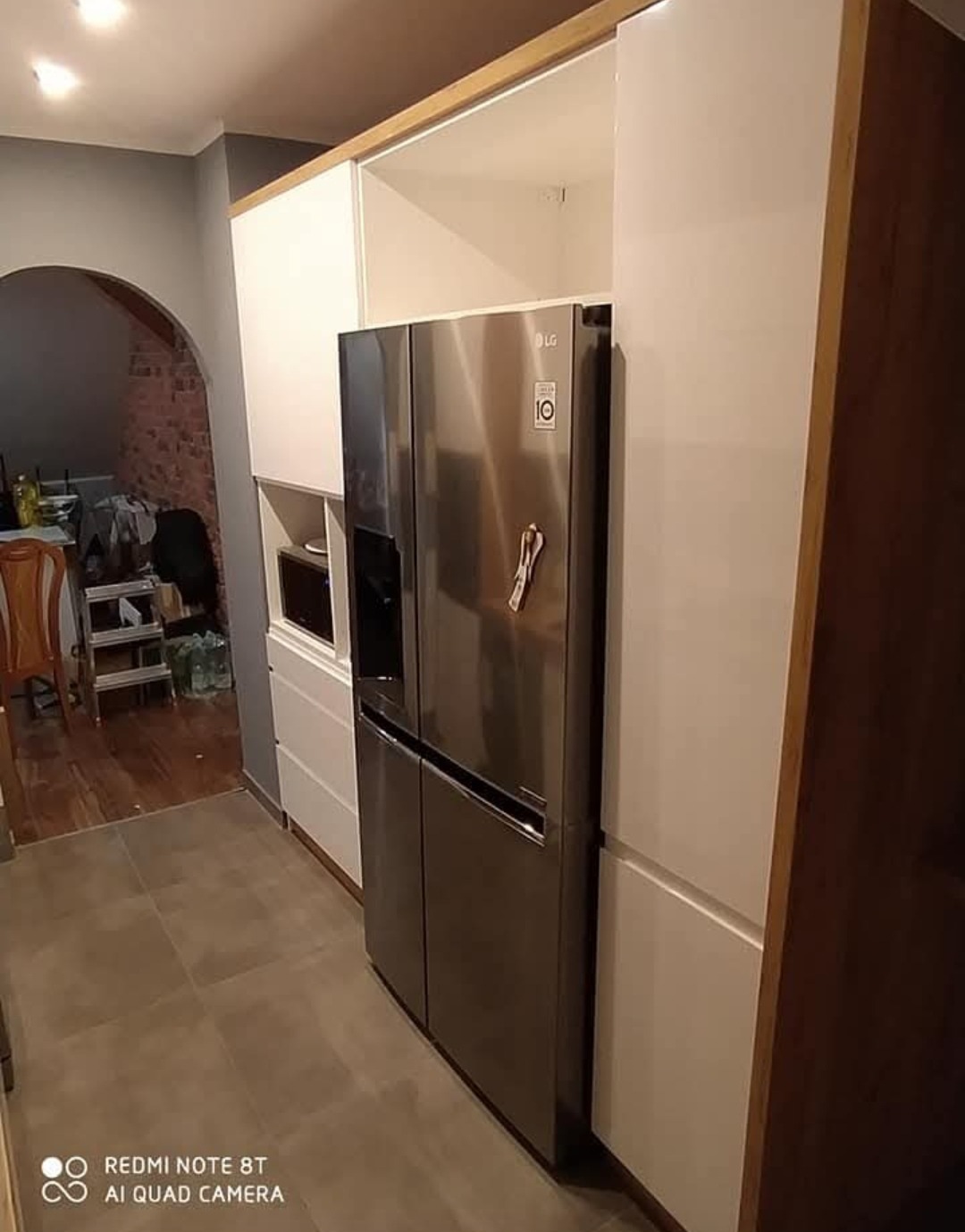 Nowoczesna kuchnia z białymi szafkami i drewnianymi akcentami, zabudowana lodówka LG, szare płytki podłogowe, widok z boku, łukowe przejście do innego pomieszczenia.