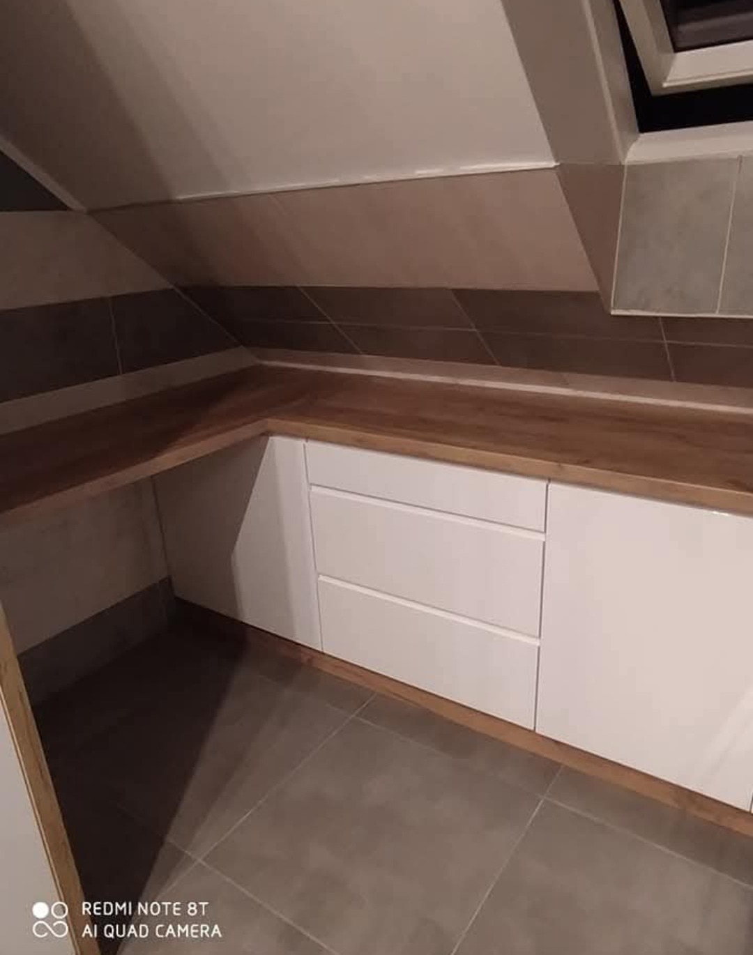 Biała szafa wnękowa z drewnianym blatem, dopasowana do skosu poddasza. Widoczne trzy szuflady i płytki na ścianie w odcieniach brązu i beżu.