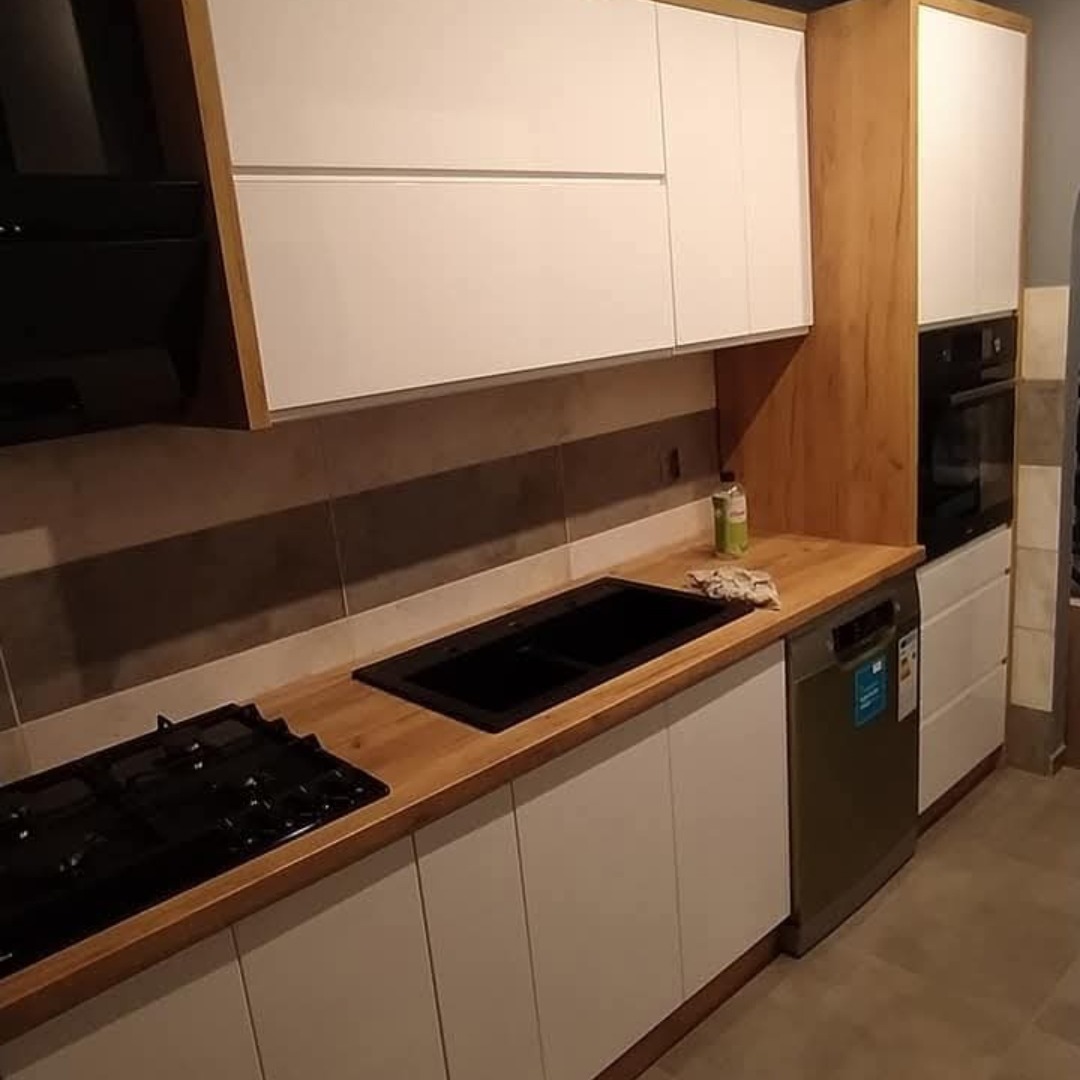 Nowoczesna kuchnia na wymiar w Radomsku: białe fronty szafek, drewniany blat i akcenty, czarny zlew i płyta gazowa. Minimalistyczny design, funkcjonalność.
