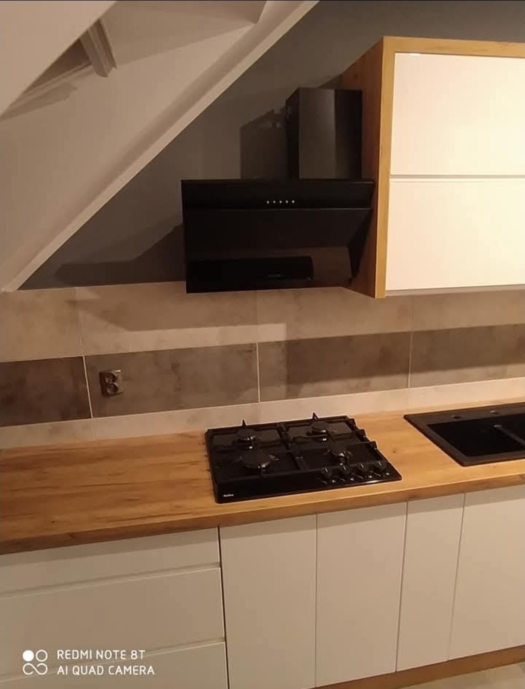 Nowoczesna kuchnia: białe szafki z drewnianym blatem i czarną płytą grzewczą. Uwagę zwraca skośny sufit i dopasowany okap.