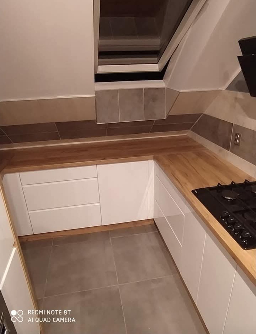 Biała kuchnia na poddaszu z drewnianym blatem i oknem dachowym. Ułożone płytki na ścianie tworzą pas. Płyta gazowa wbudowana w blat.