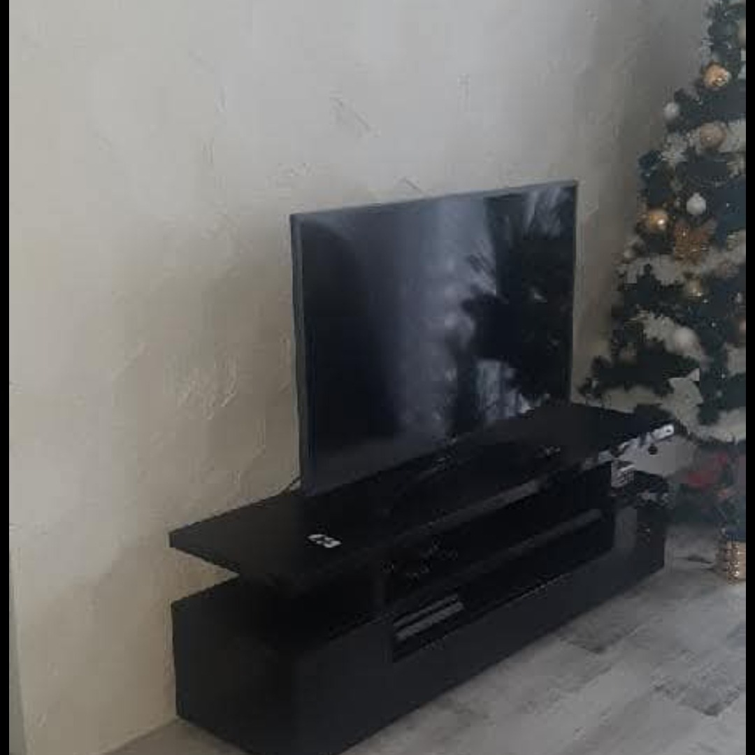 Czarna szafka RTV z telewizorem, minimalistyczny design, obok choinka. Widok z boku, jasne tło, nowoczesne wnętrze.